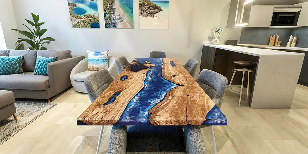 A stunning river-style resin and wood dining table with a vivid ocean-blue epoxy center, placed in a modern coastal-themed dining room with grey upholstered chairs and beach-themed wall art.