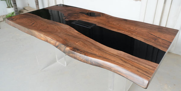 A live-edge wooden desk with a rich dark brown grain pattern and a glossy black resin inlay channel, supported by clear acrylic legs.