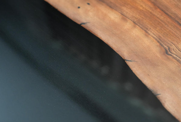 Close-up of a live-edge wooden tabletop with a rich warm brown grain and natural bark edge, set against a dark reflective surface.