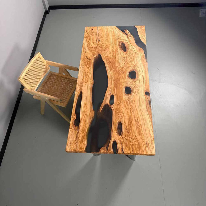 Wooden table with black resin inclusions and a wicker chair on a gray floor.