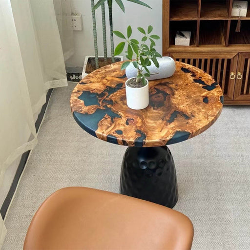 Round burl wood and black resin side table with sculpted base, styled in a cozy modern room with plants and soft lighting.
