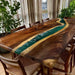 Wooden dining table with green epoxy resin inlay in a room with plants and curtains.
