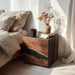 Wooden nightstand with a vase and mug next to a bed in a softly lit room