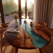 Dining table with a unique design featuring a wooden top and a blue resin river, set in a bright room with large windows.