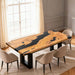 Dining table with black and wood design in a room with large windows and beige chairs.