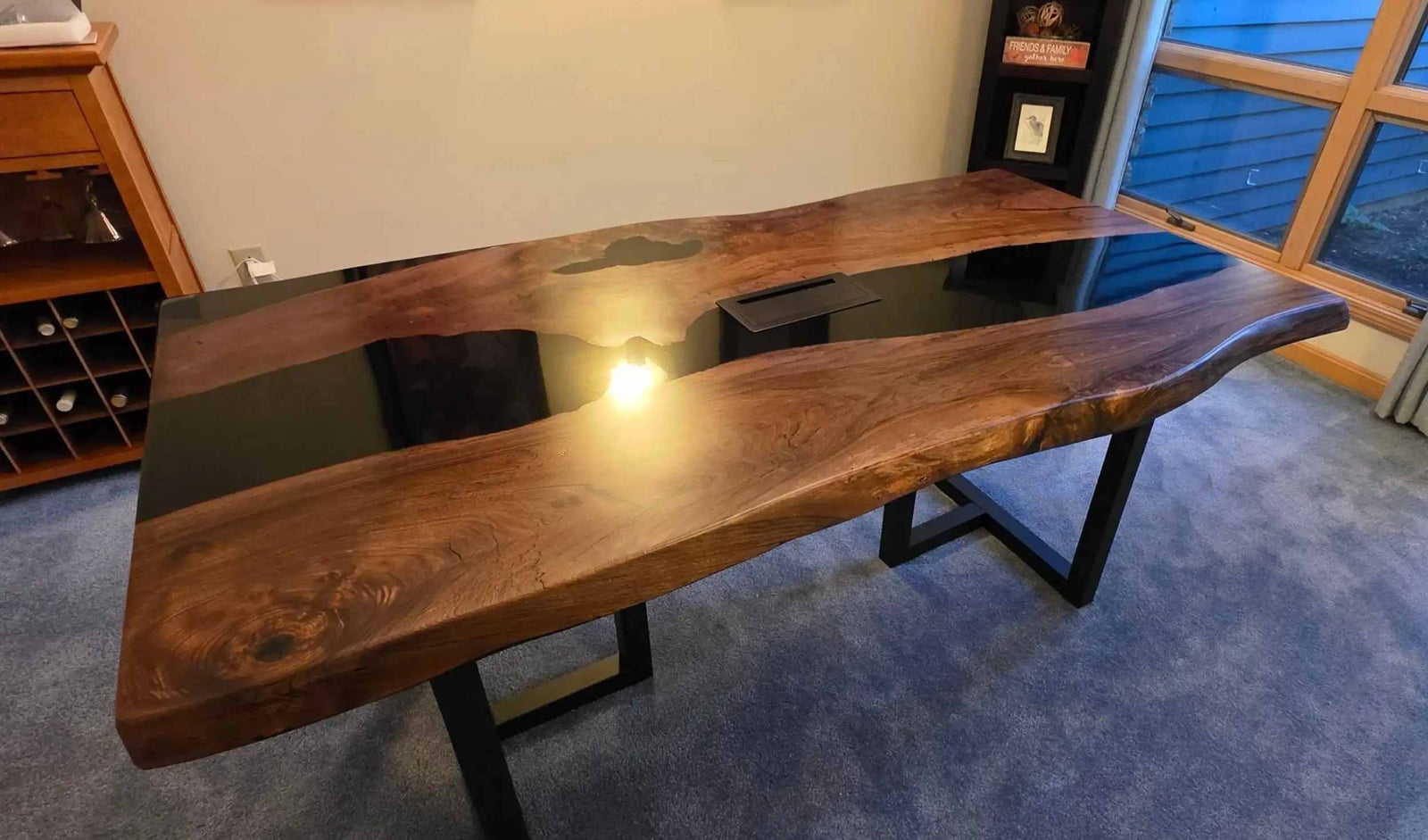 A large live-edge wooden desk with a glossy black resin river running through the center, supported by black metal legs, photographed in an indoor room with blue carpet and a window in the background.