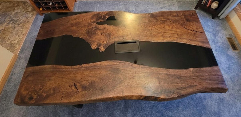 A rustic live-edge wooden table with a glossy black resin inlay running down the center, photographed from an overhead angle in an indoor room with carpet and furniture partially visible.