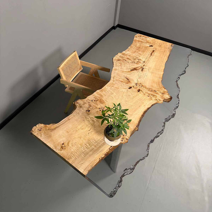 Wooden dining table with a natural edge design, accompanied by a chair and a plant on a gray floor.