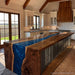 A large kitchen island with a thick live-edge wood countertop and a vivid blue river-style inlay, set in a bright modern kitchen with white cabinets, wood beams, and a window overlooking a field.