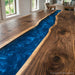 A close-up of a live-edge wooden table featuring a vibrant, swirling blue resin channel running down the center, with warm brown wood grain visible on both sides. The table is set in a bright, modern dining room with white brick walls and large windows.