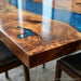 Close-up of a live-edge wooden table with a glossy finish and a swirling blue resin inlay, with parts of dark chairs visible underneath.
