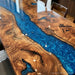 A close-up of a polished live-edge wooden table featuring dramatic swirling natural wood grain in warm brown and tan tones, divided by a vivid blue resin channel.
