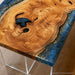 A close-up of a live-edge wooden table with a richly textured natural wood grain top, dark blue resin details, and gold leaf accents, supported by clear acrylic legs.