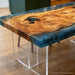 A live-edge wooden table with a richly grained natural wood top, a glossy blue resin edge, and scattered gold glitter, supported by clear acrylic legs in an indoor setting.
