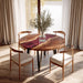 Round wooden dining table with four chairs in a bright room with large windows and curtains.
