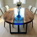 A masterful handcrafted oval black walnut and blue swirl resin river table serving as the stunning centerpiece in a high-rise dining room with floor-to-ceiling city views.