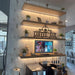 Coffee bar setup with a television screen displaying menu options, surrounded by shelves with plants and decor.