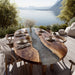 Wooden dining table with a natural edge design, set for a meal with chairs around it, on a deck overlooking a lake.