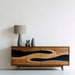 Wooden sideboard with wavy design, lamp, and decorative items on a white wall.