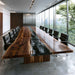 A modern conference room featuring a long live-edge wooden table with a glossy black resin river running down the center, surrounded by black office chairs and large floor-to-ceiling windows overlooking a green forest.