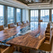 A large modern conference table with a live-edge natural wood top and a glossy dark resin river inlay, surrounded by brown leather chairs in a high-rise office with floor-to-ceiling windows.