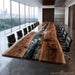 A large modern conference table made of richly graded live-edge wood with a dark river stone or resin inlay running down the center, surrounded by black office chairs in a contemporary office setting with floor-to-ceiling windows.