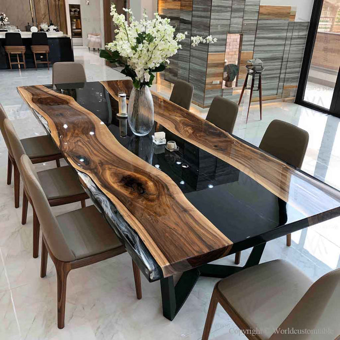 A large live-edge wooden dining table with a glossy black polished center, surrounded by modern upholstered dining chairs in a bright showroom. A vase of white flowers sits on the table as a centerpiece.