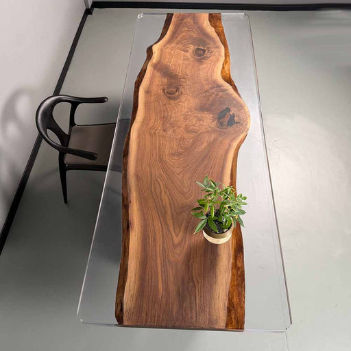 Wooden dining table with a clear glass top, featuring a potted plant on a gray floor.