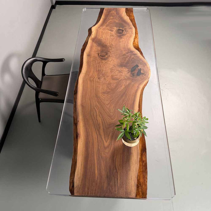 Wooden dining table with a clear glass top, featuring a potted plant on a gray floor.