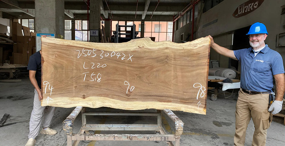 Two men holding a large wooden slab with measurements in an industrial setting.